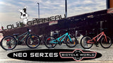 Three bicycles in different colours (dark green, teal, and coral pink) lined up in front of a brick wall with a Louis Garneau logo. A black banner at the bottom reads 'NEO SERIES BICYCLE WORLD'.